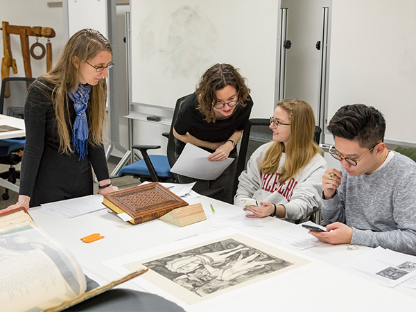 A group of students and instructors examine large prints of artwork on a drafting table.